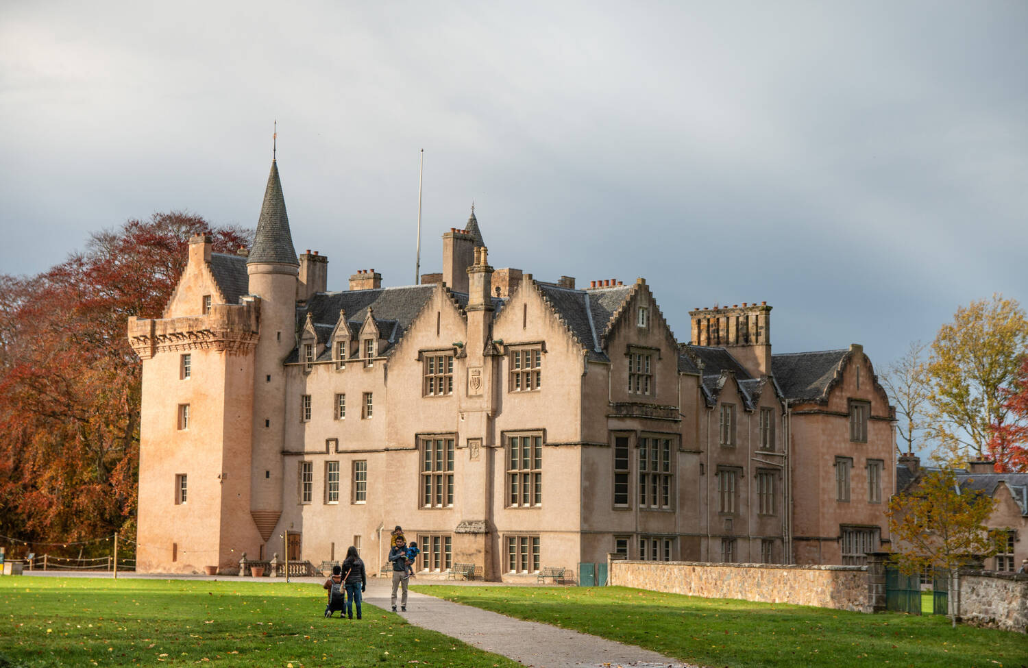 A view of the exterior of Brodie Castle on an autumn day. Tall beech trees behind the castle are red and orange. The walls of the castle have a pink hue. A couple with a pushchair walk along a gravelled path leading up to the castle.
