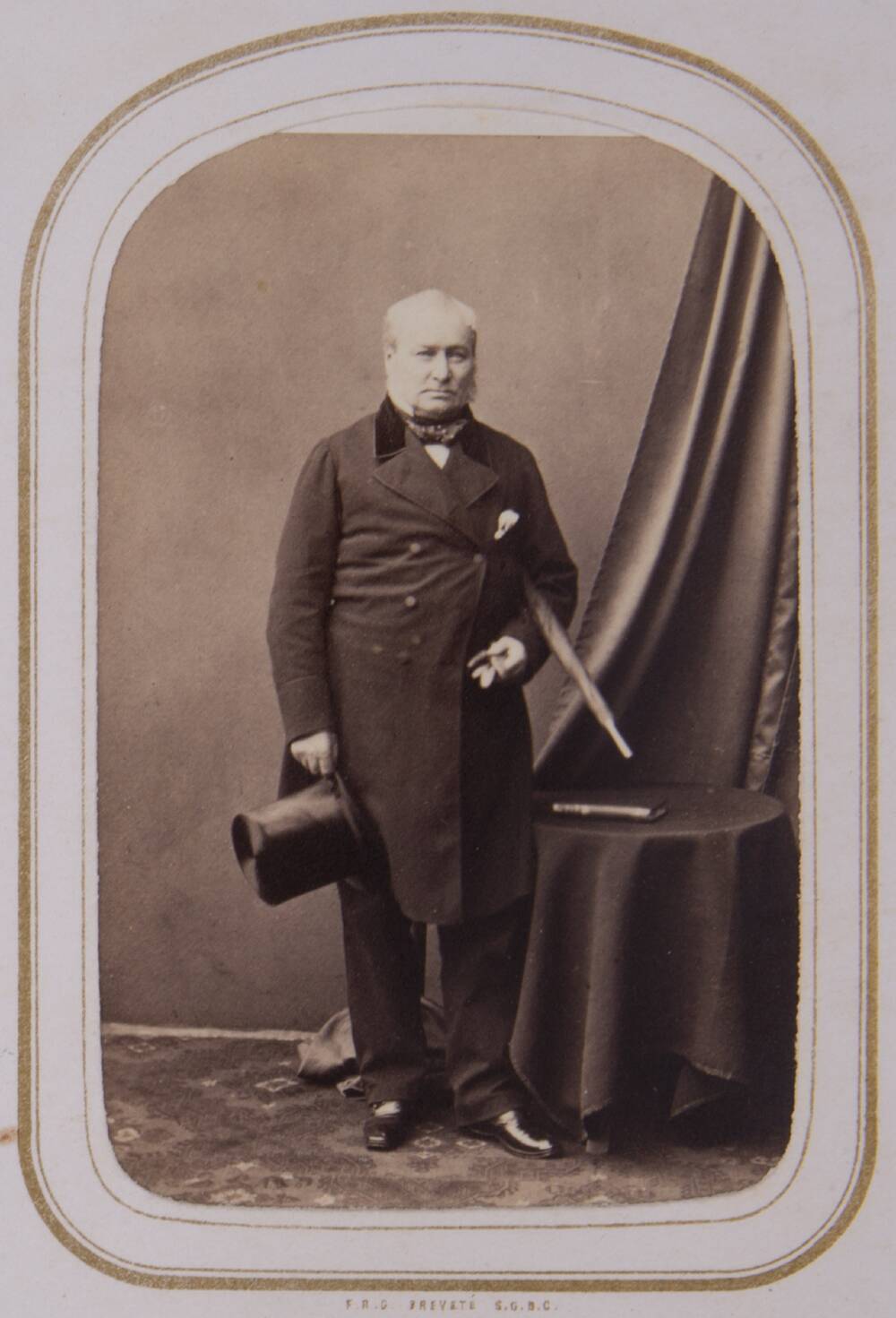 A sepia photograph of an older Victorian-looking man, standing in a photography studio. He wears a long dark overcoat and carries a top hat in one hand, with a walking cane tucked under the other arm. He stands beside a small round table covered in a black cloth.