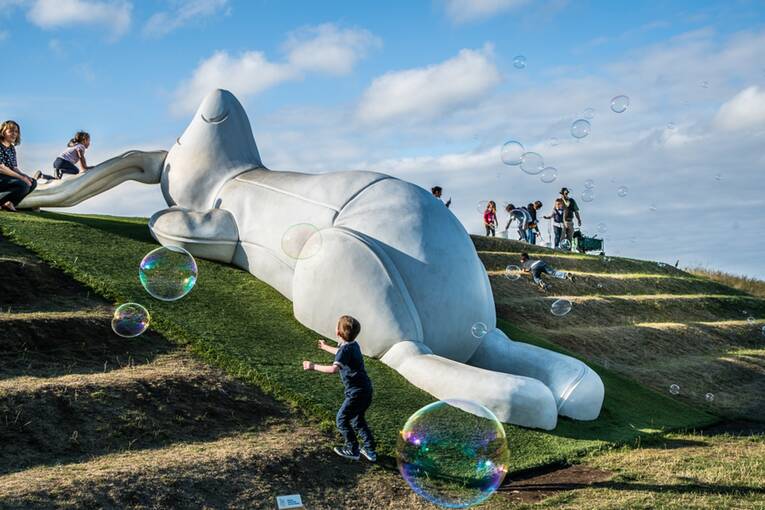 A large white statue of a rabbit lies on a hillside. Children play around it. Bubbles float in the air beside them.