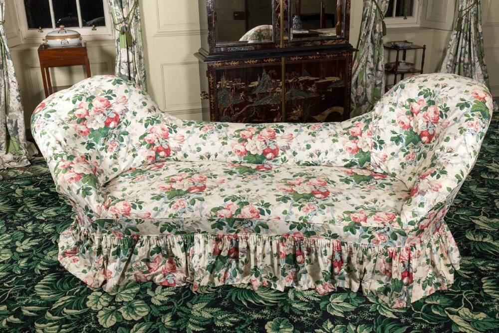 A floral-fabric sofa stands on a green, leaf-patterned carpet. The sofa looks almost like a chaise longue, as it has two raised ends with a low dip in the middle.