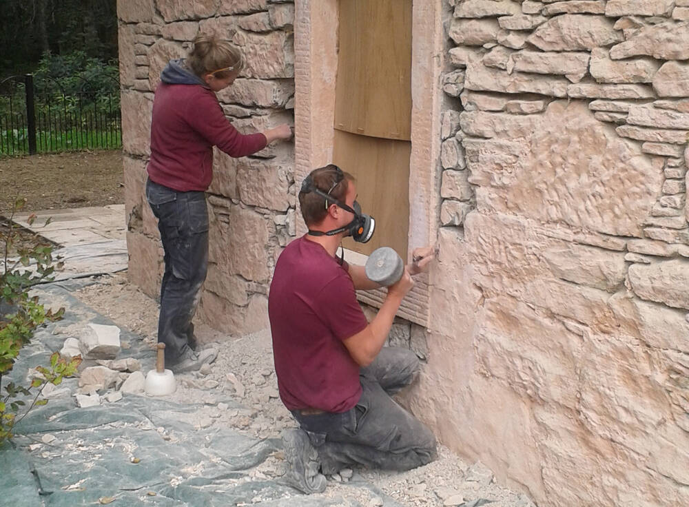Two stonemasons work on a building beside a boarded-up window. The man kneeling in the foreground wears a mask and holds a large tool.
