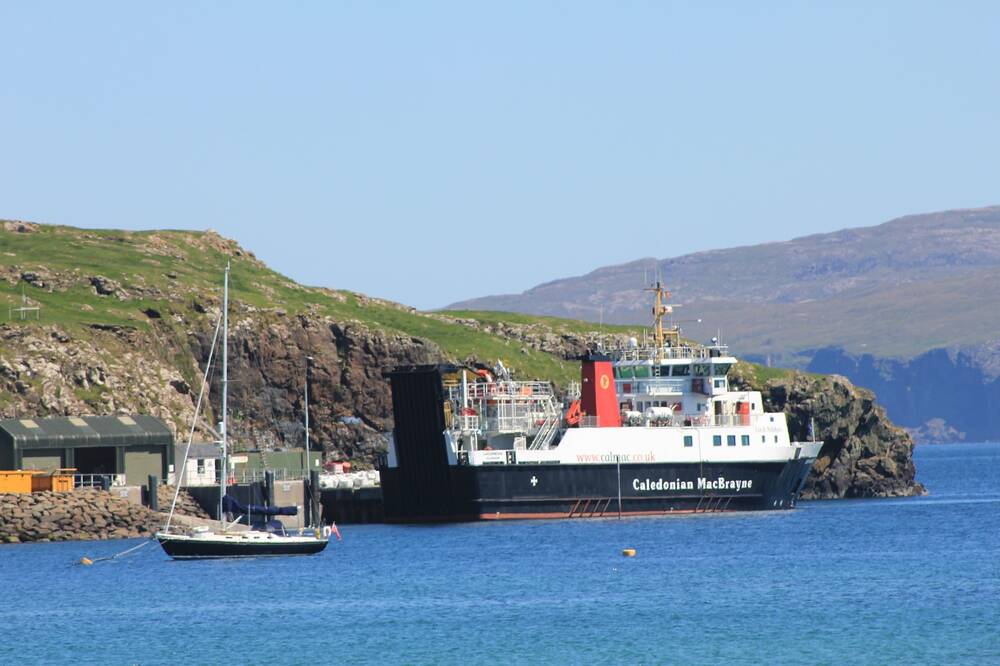 A CalMac passenger ferry is berthed alongside a pier on the Isle of Canna. A rocky slope rises behind it. A yacht is anchored out in the water in the foreground.