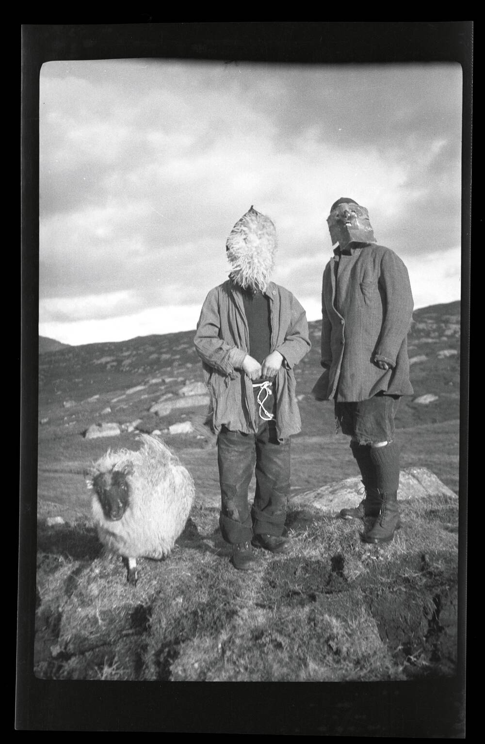 A black and white photograph of two figures standing in a boulder-strewn field, next to a large sheep. Both boys wear costumes. The one on the left wears a round piece of sheep fleece completely covering his face. The boy on the right has a crudely shaped metal mask.