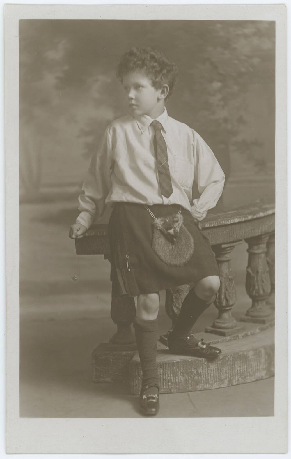 A full-length formal black and white photograph of a young boy. He stands by a banister and looks away to the left. He is wearing a white shirt, tartan tie, kilt, sporran and woollen knee-high socks.