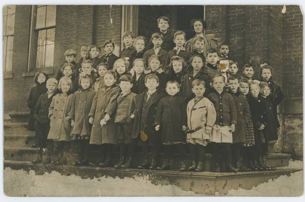 A group of around 40 schoolchildren (girls and boys aged between 5 and 10) stand on some steps outside a stone building. Their teacher stands on the top step. Deep snow lies at the base of the steps and the children all wear boots and coats.