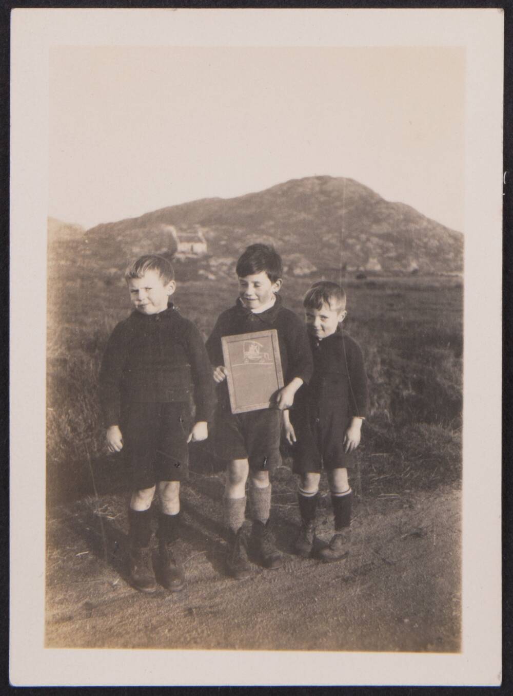 A black and white photograph of three young schoolboys standing at the edge of a road. Hills and a single cottage stand in the background. The boys all wear black jumpers with dark shorts. The boy in the middle is holding a slate with a chalk drawing on it.