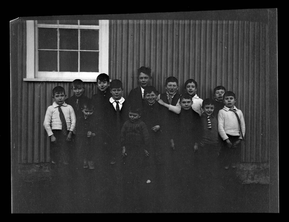 A black and white photograph of a group of 14 schoolboys (of mixed ages) standing in front of a corrugated schoolhouse and a white-framed window. Most of the boys are wearing a tie.