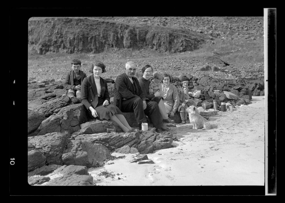 A black and white photograph of a group of people sitting on some rocks on a beach. A tin mug is balanced on the rock in the foreground. A small dog sits on the sand and looks up at a young boy, who is sitting on the knee of a young woman.