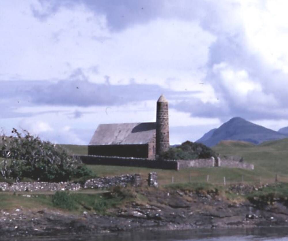 An old colour photo of a church on an island. It has a spire shaped a little like a rocket. In the background is a conical mountain.