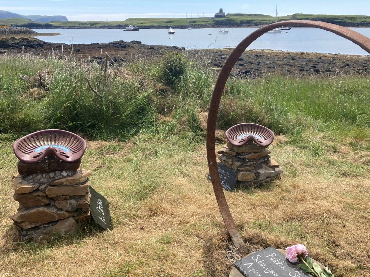 Stone cairns with old tractor seats attached to the top stand beside each other. Slates lie against the cairns, with names painted on it. Across the bay, in the distance, can be seen St Edward's Church, pictured through a large iron cartwheel hoop.