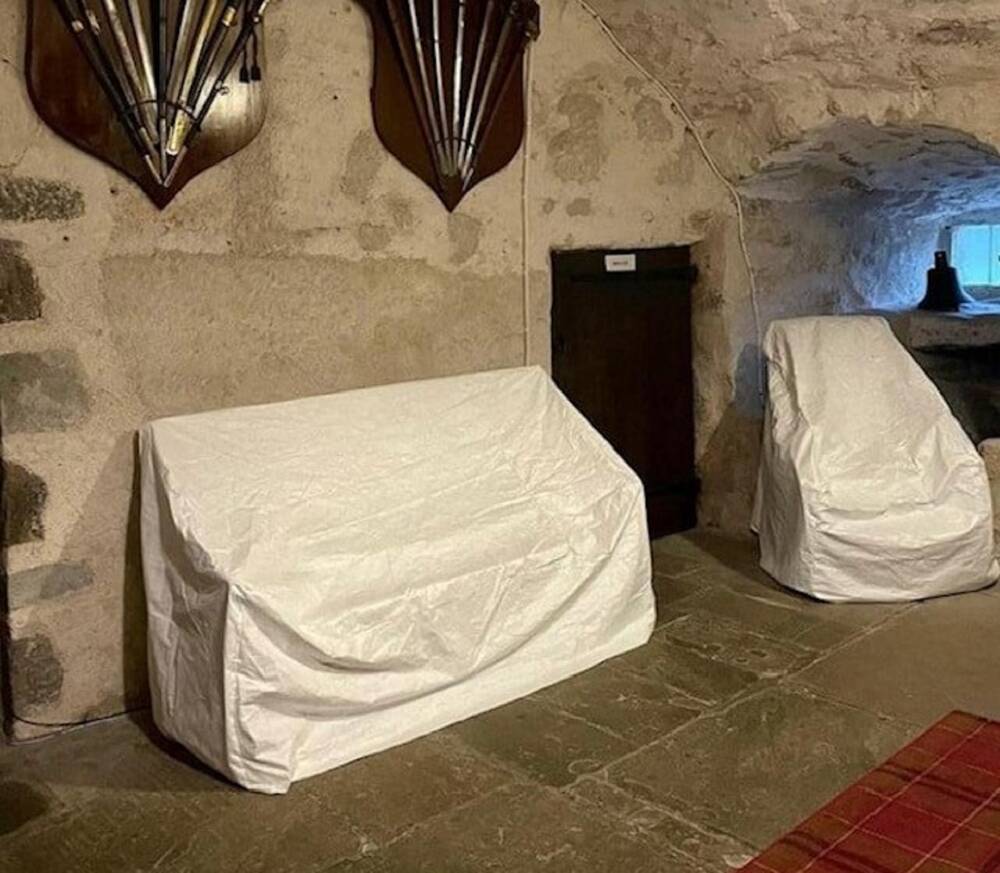 Two large collection items are covered in white sheets in a large stone hall of a castle. Two wooden shields hang on the stone wall above them.