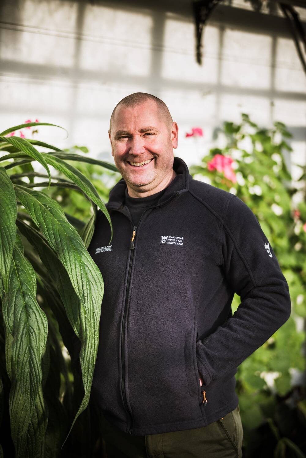 A smiling man in a navy National Trust for Scotland fleece stands inside a glasshouse, surrounded by plants.