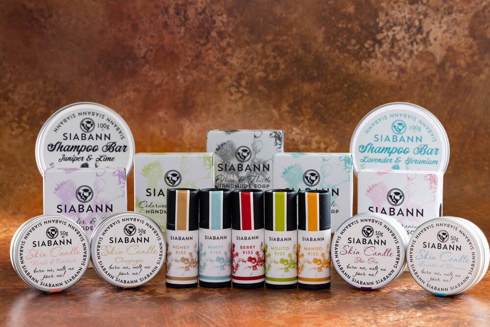 A range of Siabann beauty products are displayed against a brown marbled background. At the front are small tubes of lip balm and little candles in tins. Behind are soap boxes and tins of shampoo bars.