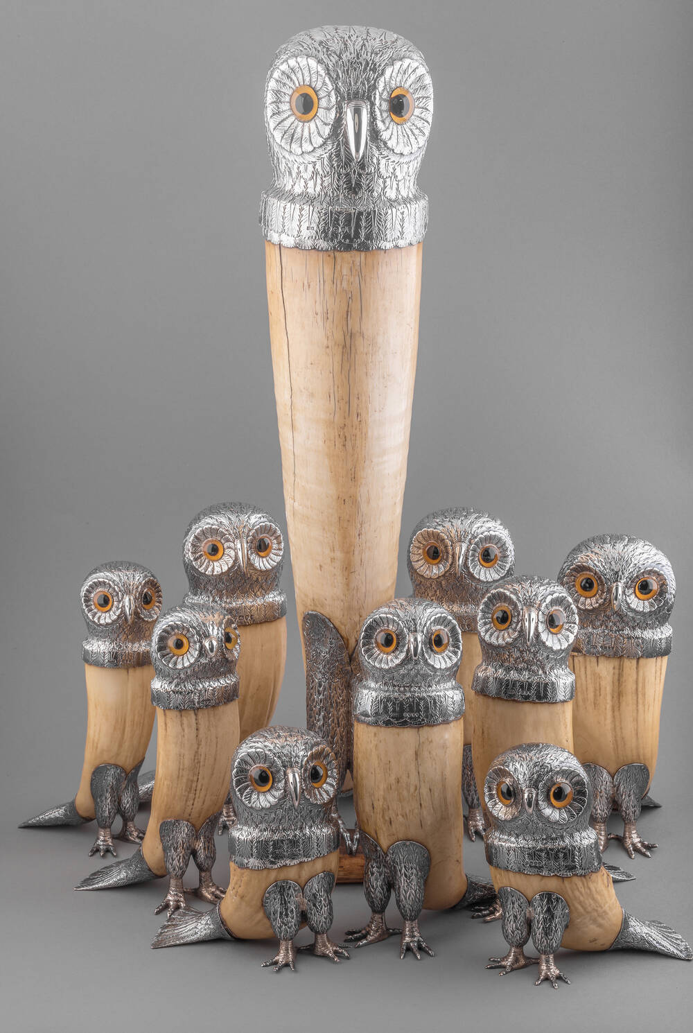 A group of 19th-century owl-shaped drinking horns, with 9 small owls and 1 very tall one.The bases are made from a light-brown horn, and all have a silver owl head on top, with orange eyes.