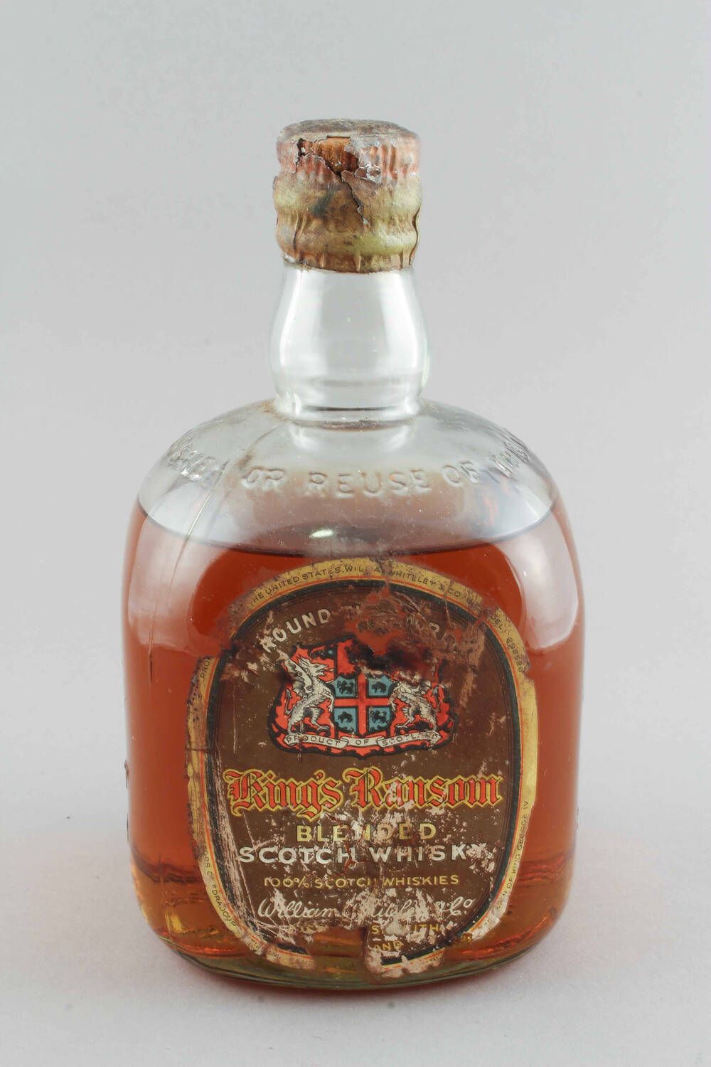 An old bottle of King’s Ransom whisky. The whisky is orange in colour and can be seen through the clear glass bottle. The label has worn away in places and the seal over the cork has split a little.