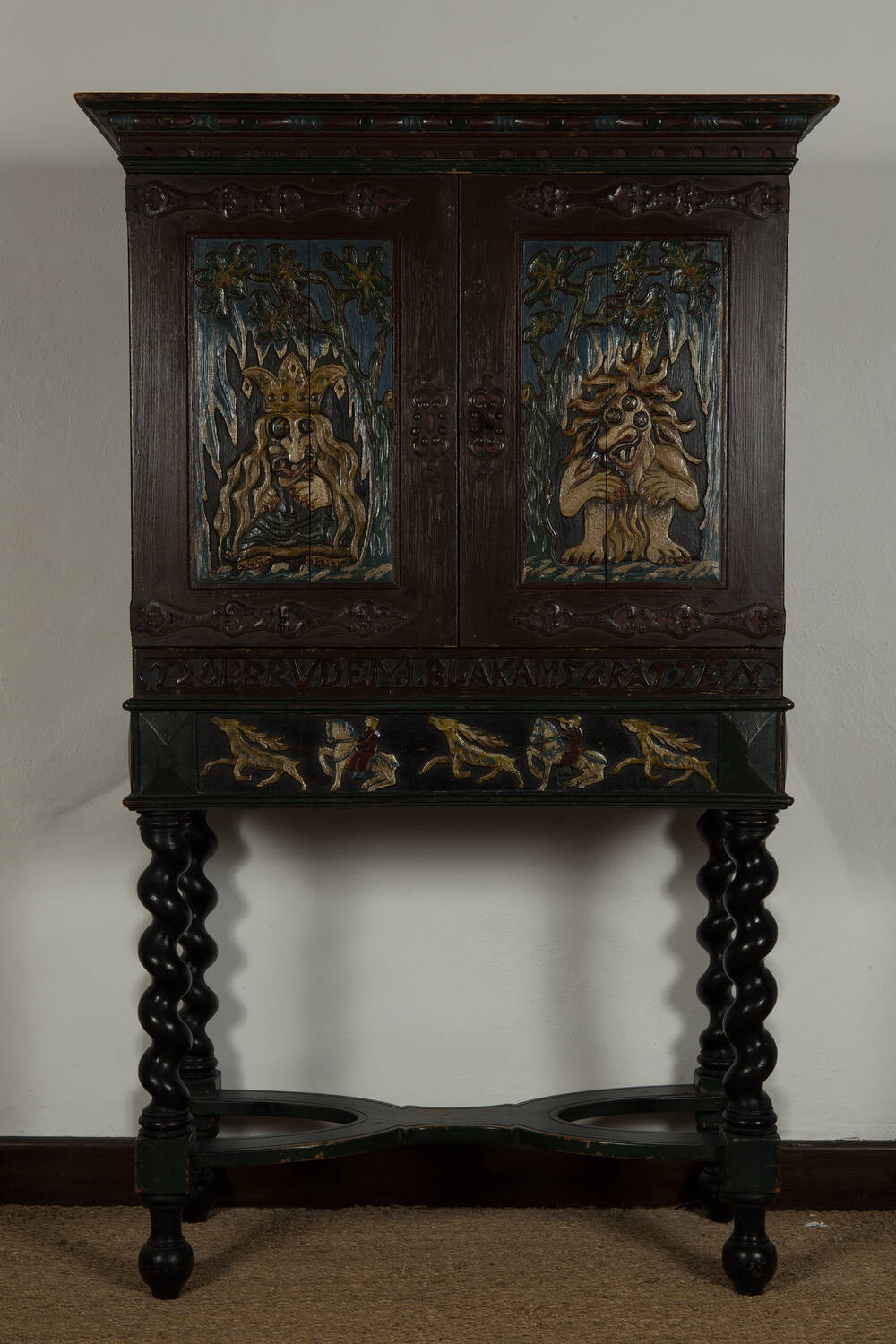 A tall, dark wooden cabinet with spindle legs. The doors are adorned with carvings of trolls, and carvings of horses run along the bottom of the cabinet.