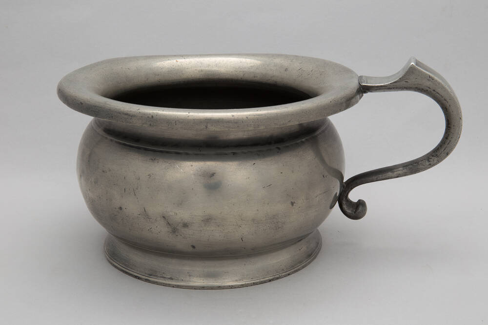 A pewter chamber pot with a handle to the right.