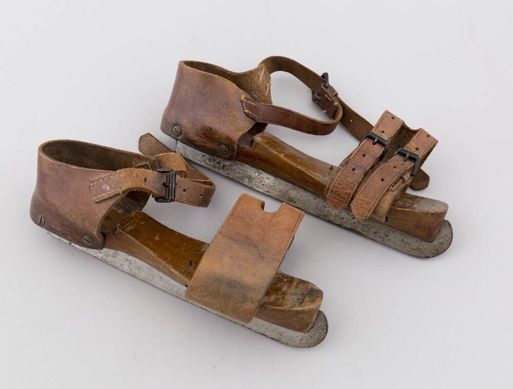 A pair of old ice skates, with a wooden base, metal blades and leather upper straps.