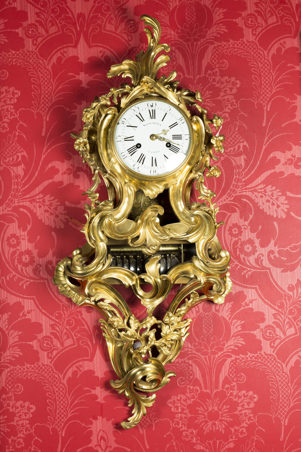 A very ornate golden cartel clock, hanging on a wall covered with red paper.