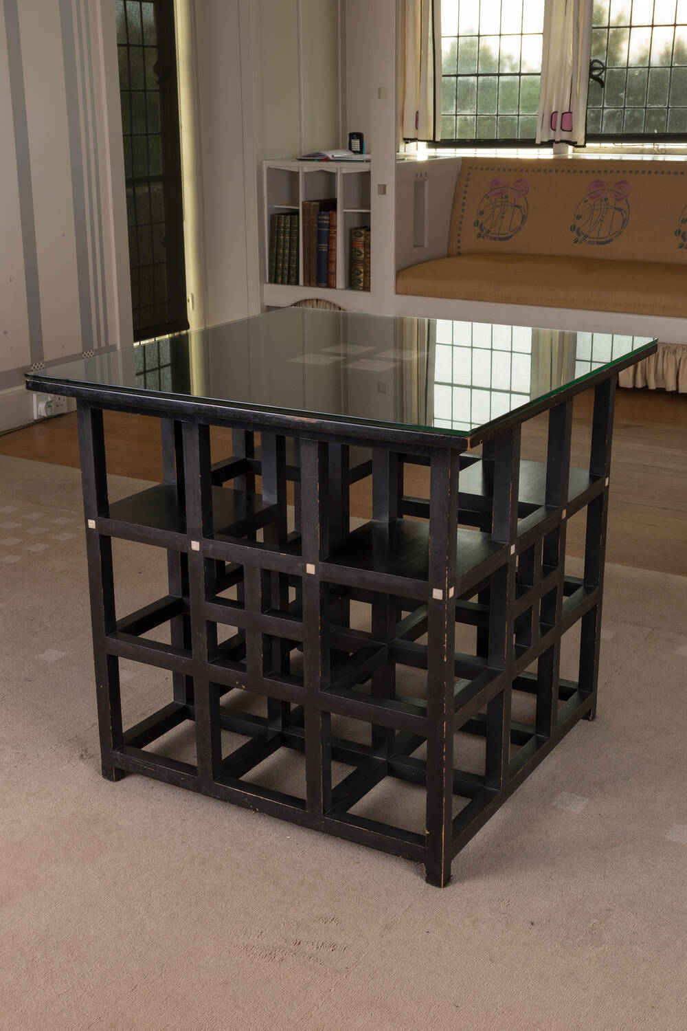 A wooden centre table designed by Charles Rennie Mackintosh. The top is inlaid with mother-of-pearl and covered by a glass square. The base is made up of a grid of wooden squares.