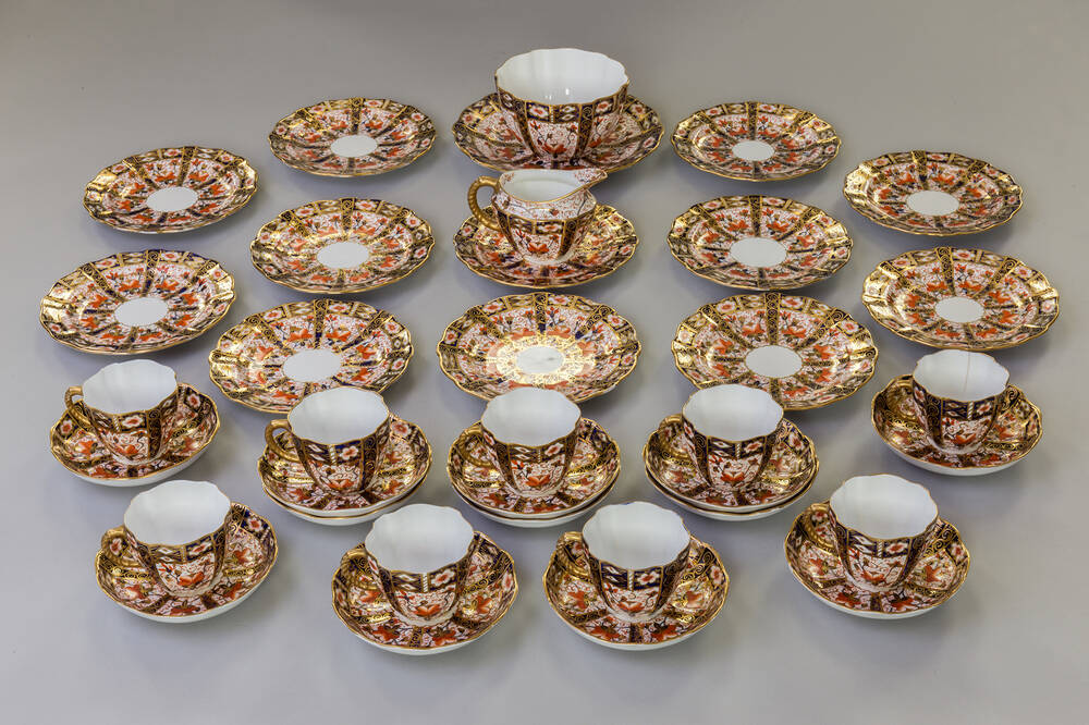 A fine Royal Crown Derby porcelain tea set is all laid out on display. There are tea plates, cups and saucers, a milk jug and a sugar bowl. The design is a gold, red and blue pattern.