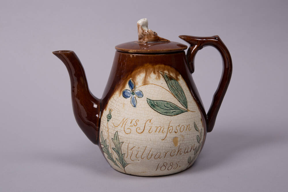 A varnished ceramic teapot with a brown spout and handle. The body has leaves and flowers painted on it, as well as the words: Mrs Simpson, Kilbarchan 1885.