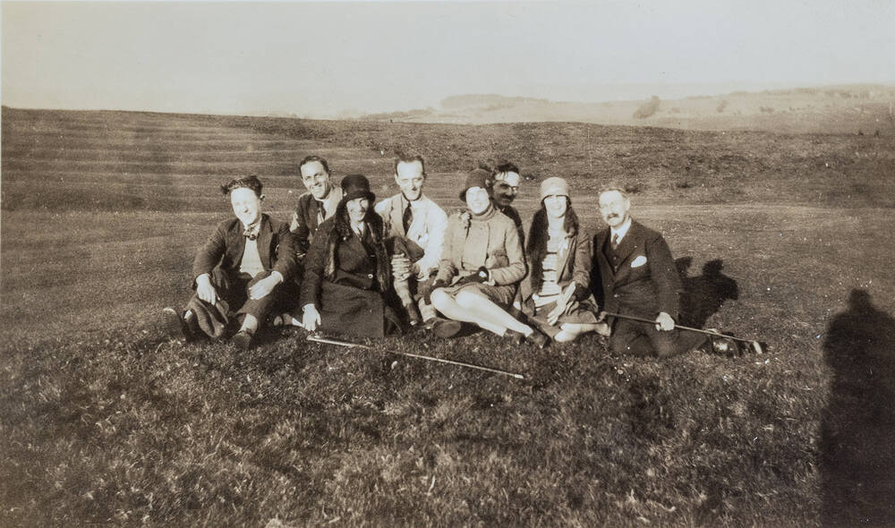 A black and white photo of a group of men and women sitting on a golf course