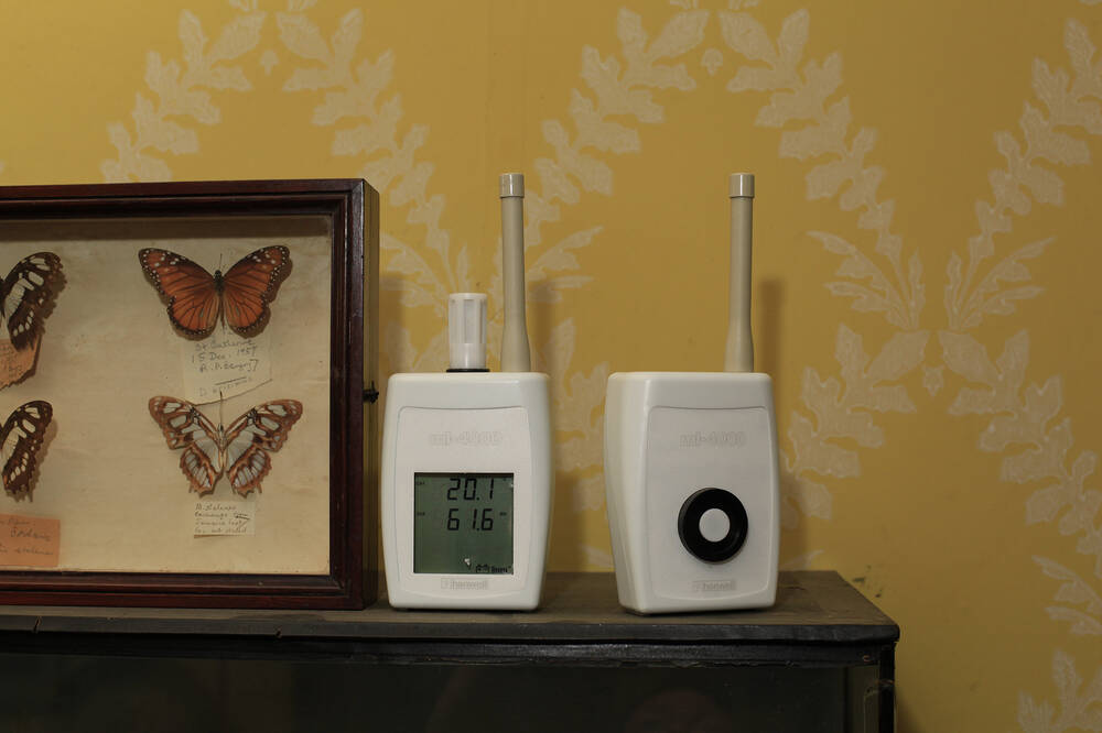 Two environmental monitors sit on a shelf beside some framed butterflies, in Canna House. The wallpaper in the background is yellow with a white leaf pattern.