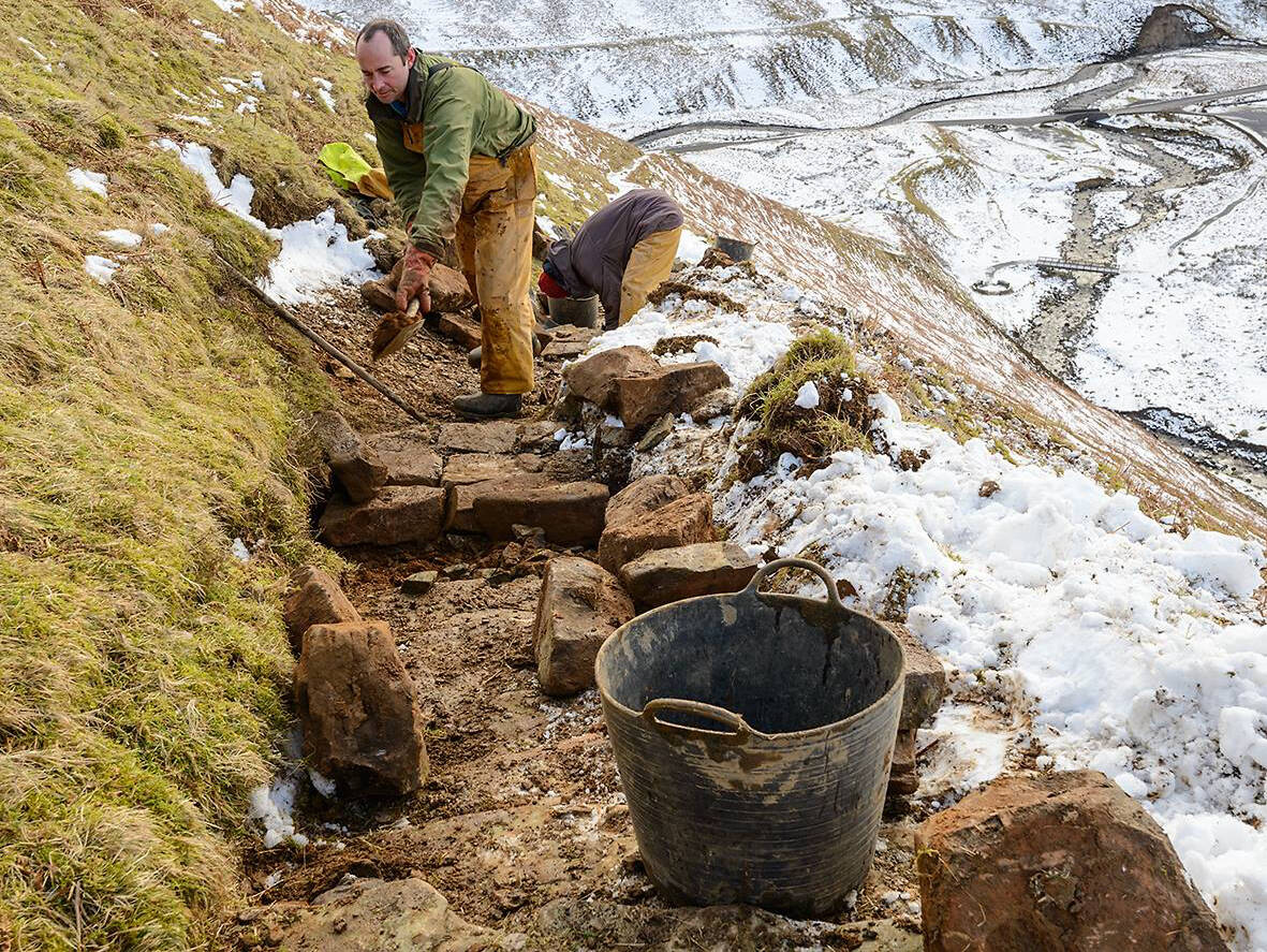 A small group of people work at repairing a mountain path. The man in the foreground is digging a space to add large rocks. There is snow on the hillside surrounding the narrow path.