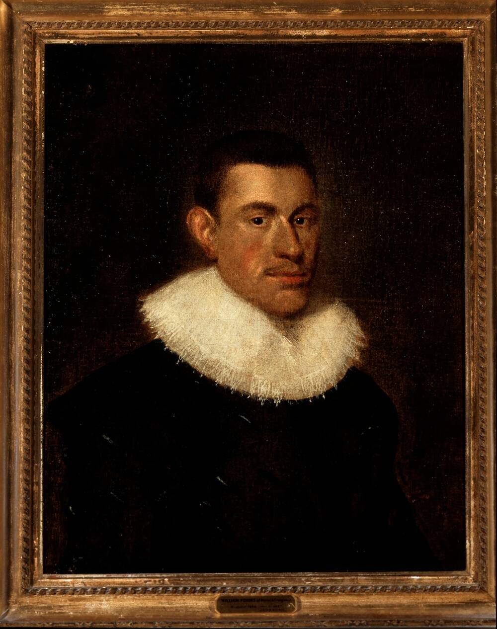 A framed portrait of a man against a dark plain background, wearing black clothes and a wide white ruff high on his neck. He is looking directly out towards to the viewer: his head is turned slightly to the side but his eyes are forward.