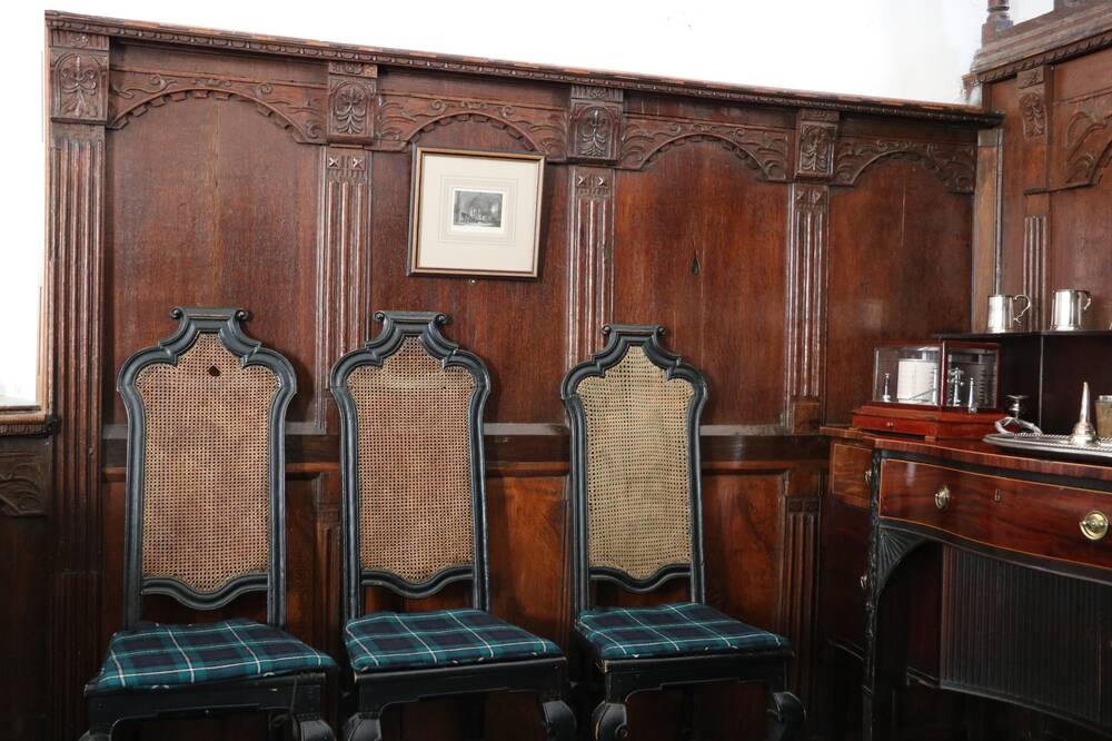 Three high-backed dining chairs with tartan seats sits against a wood-panelled wall. The wood is carved and features columns as well as detailing along the top. At the corner of the room, the wood continues along the next wall.