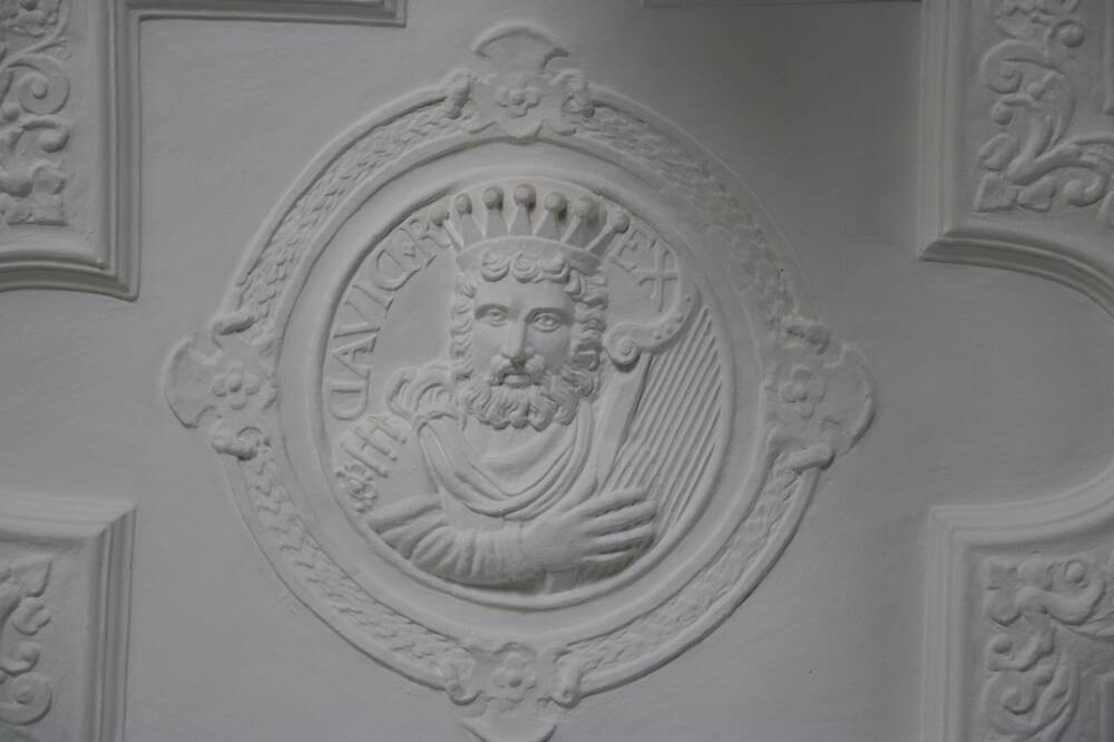 A section of white plaster ceiling, adorned with the moulding of the head and shoulders of a crowned and bearded king holding a lyre. Beside his head is the word ‘David’ in reverse.