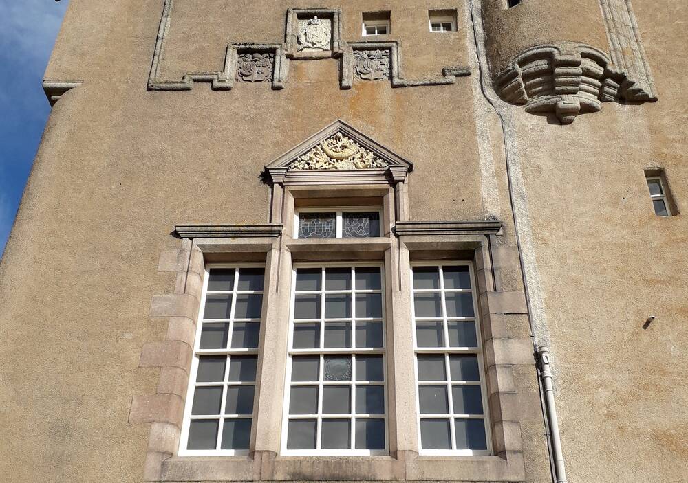 A close-up view of the three stone armorial panels on the castle wall, above a triangular decorated stone, which is above a large window.