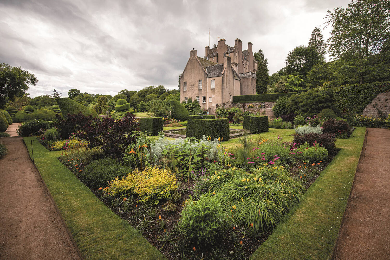 A view of the walled garden at Crathes Castle, looking towards the castle diagonally across a square flower bed. Shaped yew hedges surround a square pool at the centre of the bed, with colourful flower beds around the border. Gravel paths run along the outside.