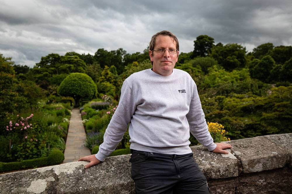 A man in a light grey sweatshirt stands against a waist-high stone wall, with Crathes Castle walled garden behind him.