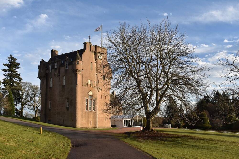 A view looking across to Crathes Castle on a sunny winter day from the parkland area. A bare horse chestnut grows in front of the castle. Behind the castle stands a very tall fir tree.