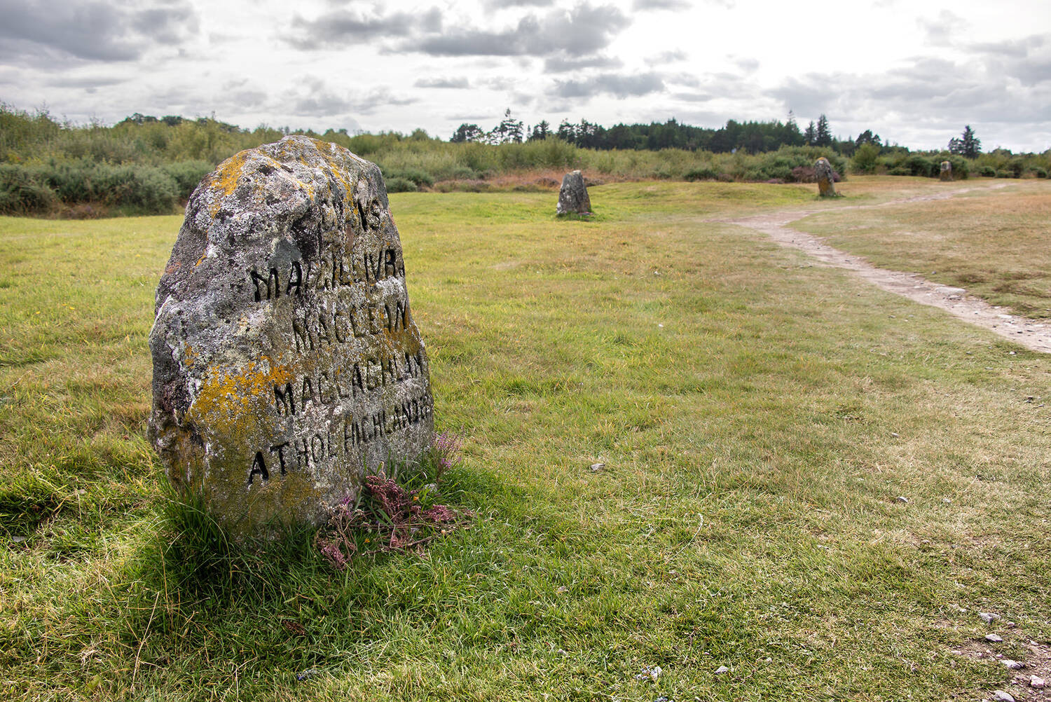 A gravestone stands beside a path on Culloden moor. The stone has lichen growing on it.