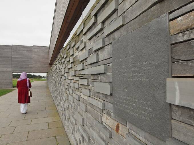 An exterior wall of Culloden Visitor Centre showing protruding grey stones from a grey wall. A plaque is inlaid to the wall. A lady walks along the path beside the wall.