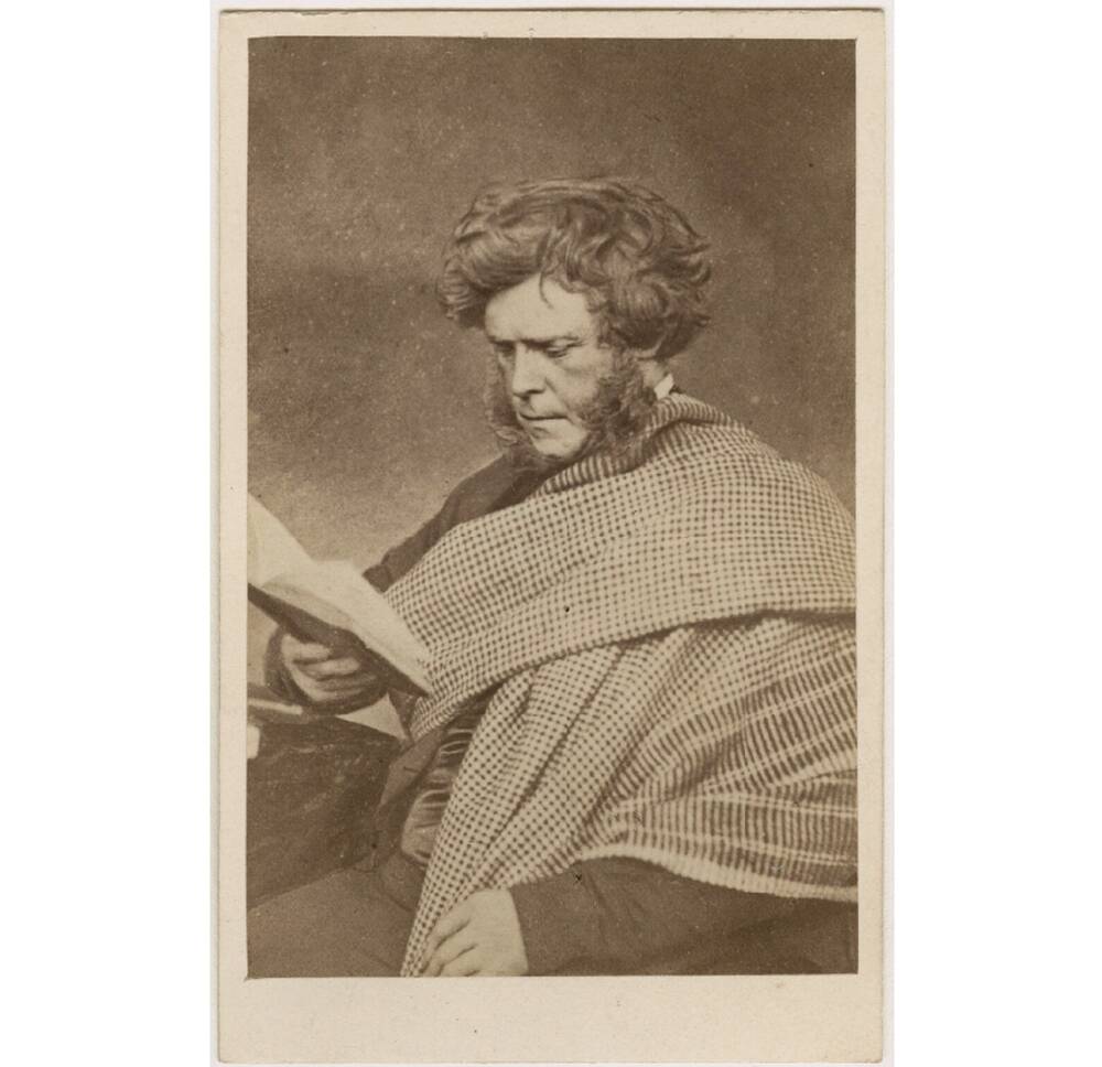 A sepia-toned photograph of an older Victorian man, shown seated and reading the book he holds in one hand. He is wrapped in a large tartan shawl. He has thick brown hair and very long sideburns.