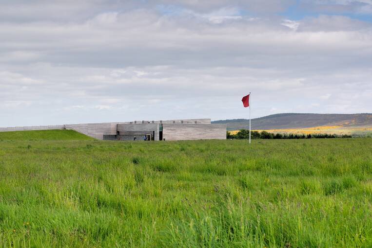 Culloden Visitor Centre can barely be seen across the battlefield, with the grey walls blending into the skyline. A tall red flag stands near the building.