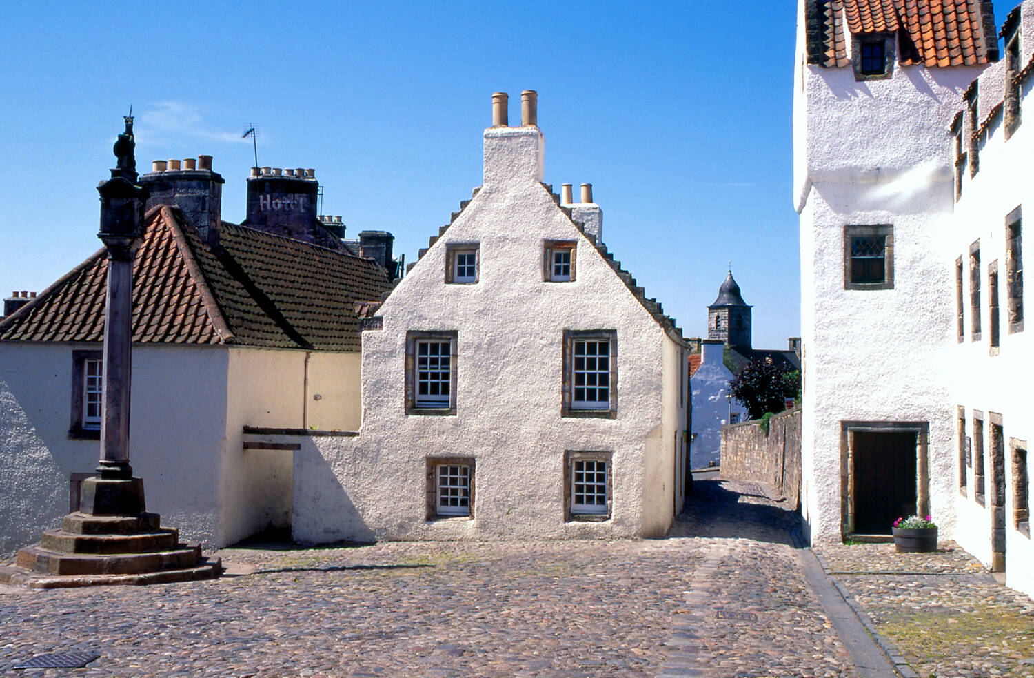 The mercat cross in the cobbled village square in Culross, surrounded by white 17th-century houses.