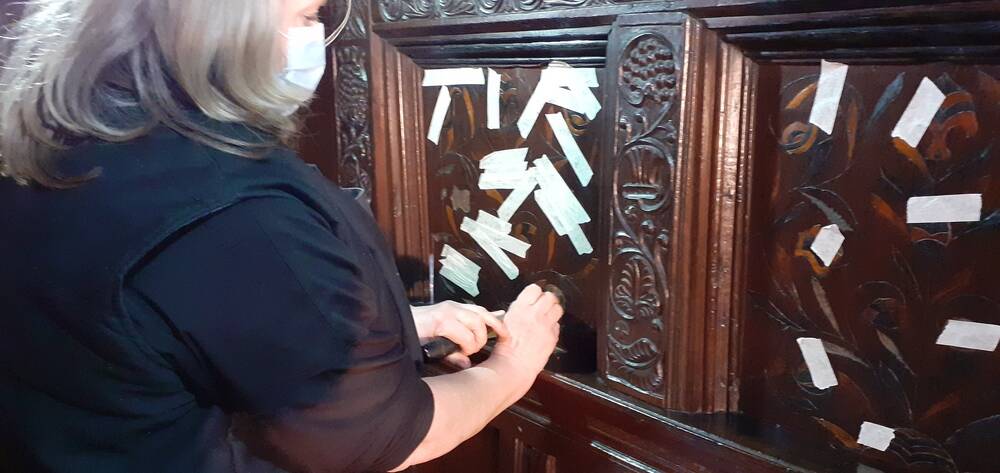 A lady works on a section of an ornately carved wooden headboard. She has taped a number of inlaid sections into place.