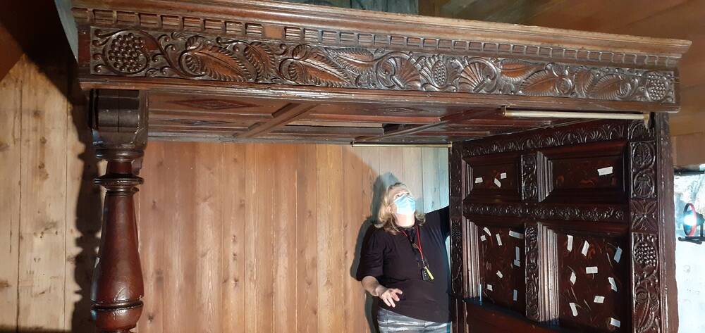 The upper section of an ornately carved wooden four-post bed in Culross Palace. In the background, a lady is standing looking up at it.