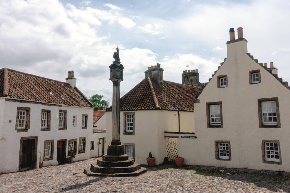 A view of the mercat cross in the centre of the village of Culross. The town square has cobblestones and is surrounded by white painted buildings with crow-stepped gables.