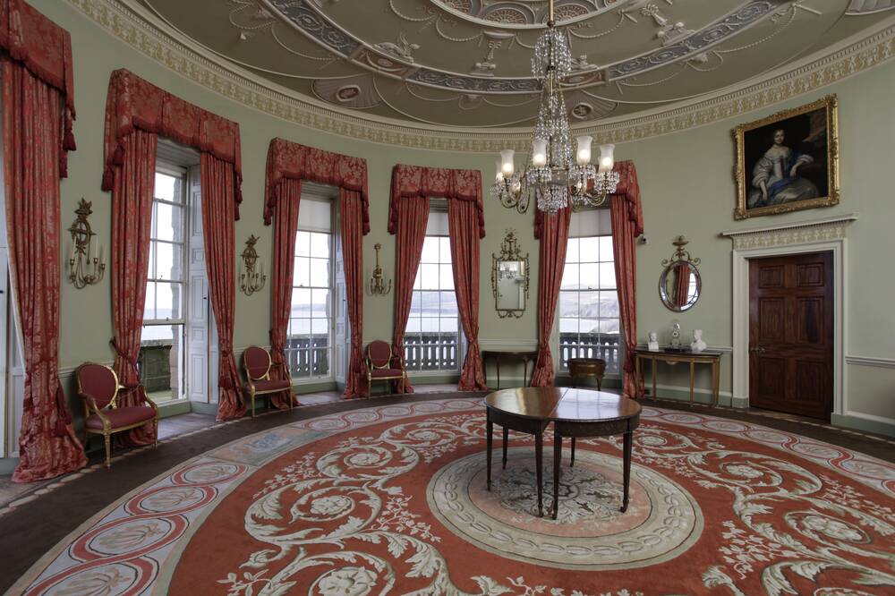 The Round Drawing Room at Culzean, with soft furnishings such as the large circular rug and curtains in a red and cream colour scheme. There are several windows overlooking the sea view and a chandelier hangs from the ceiling.