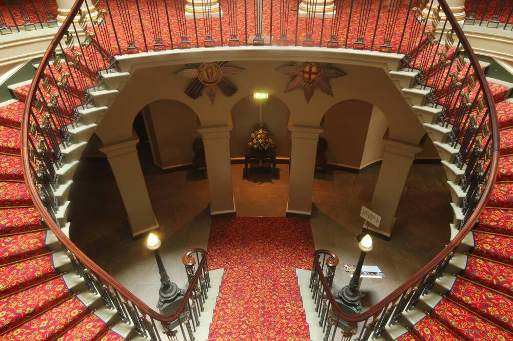 A view from above of a double staircase, swooping around an oval opening. The stairs are covered in a red and gold carpet. Two tall lamps stand either side of the bannisters at the bottom of the stairs.