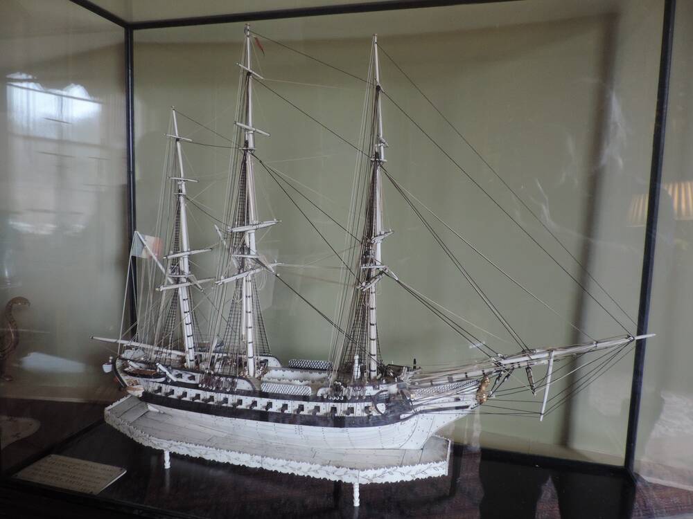 A large model of a three-masted sailing ship is displayed inside a glass cabinet. The ship is made of white bone, and stands on a matching white platform.