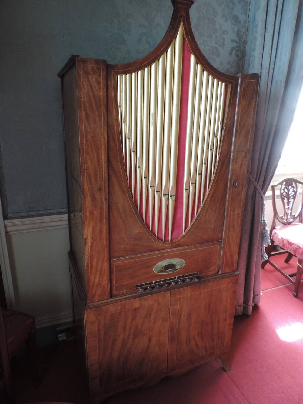 A barrel organ, housed in a tall wooden cabinet, stands by the wall, in front of blue curtains. There are silver pipes at the top of the cabinet, with a row of pegs halfway down. There are two cupboard doors at the bottom of the cabinet.