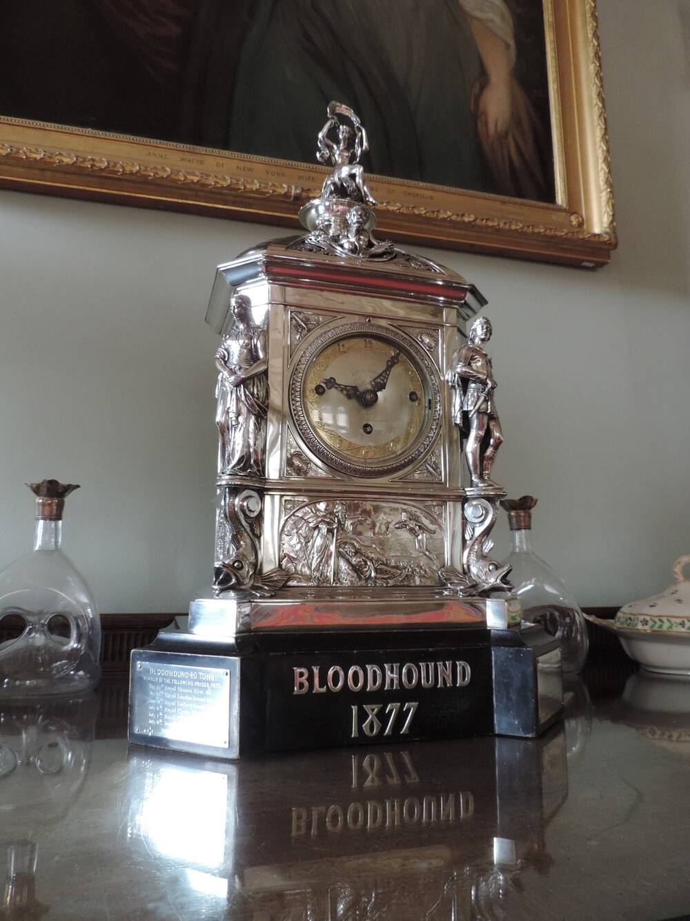 A silver carriage clock stands on a polished shelf, with two glass bottles either side. A gilt frame can be seen behind. The silver clock stands on a marble plinth, inscribed with Bloodhound 1877. There is a plaque with a list of dates on the side panel of the plinth. On top of the clock is a small silver figurine of a seated woman, holding a long banner above her head.