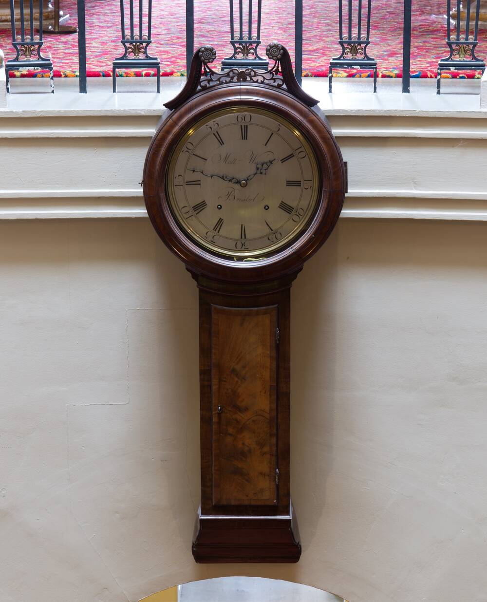 A wooden clock with the dial at the top and a long wooden tail hangs on a white wall, apparently attached to metal railings.